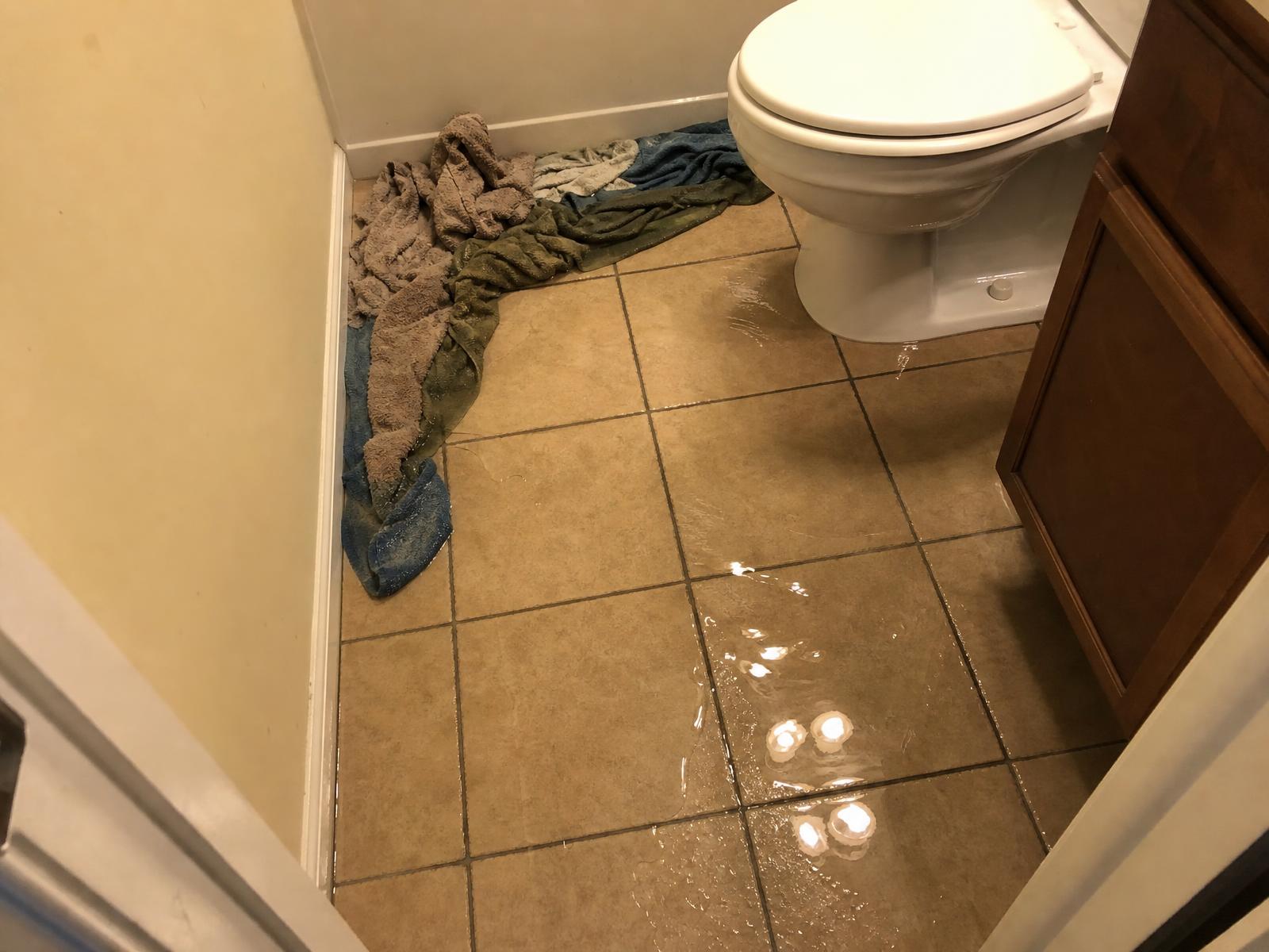 Water spreading across a tile bathroom floor from the base of a toilet with towels holding back the spill