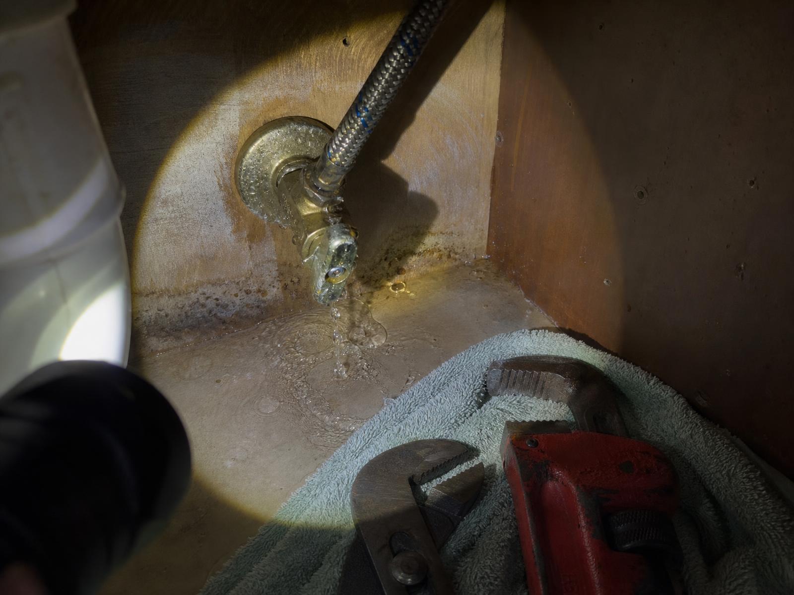 Flashlight beam on a leaking brass shutoff valve and braided supply line under a sink, with wrenches on a towel